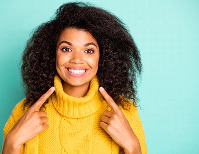 A young woman wearing a yellow sweater and pointing to her brighter smile because of teeth whitening in Ellicott City