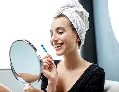 A young woman with a towel on her head, holding a syringe with whitening gel and a hand-held mirror to look at her brighter smile